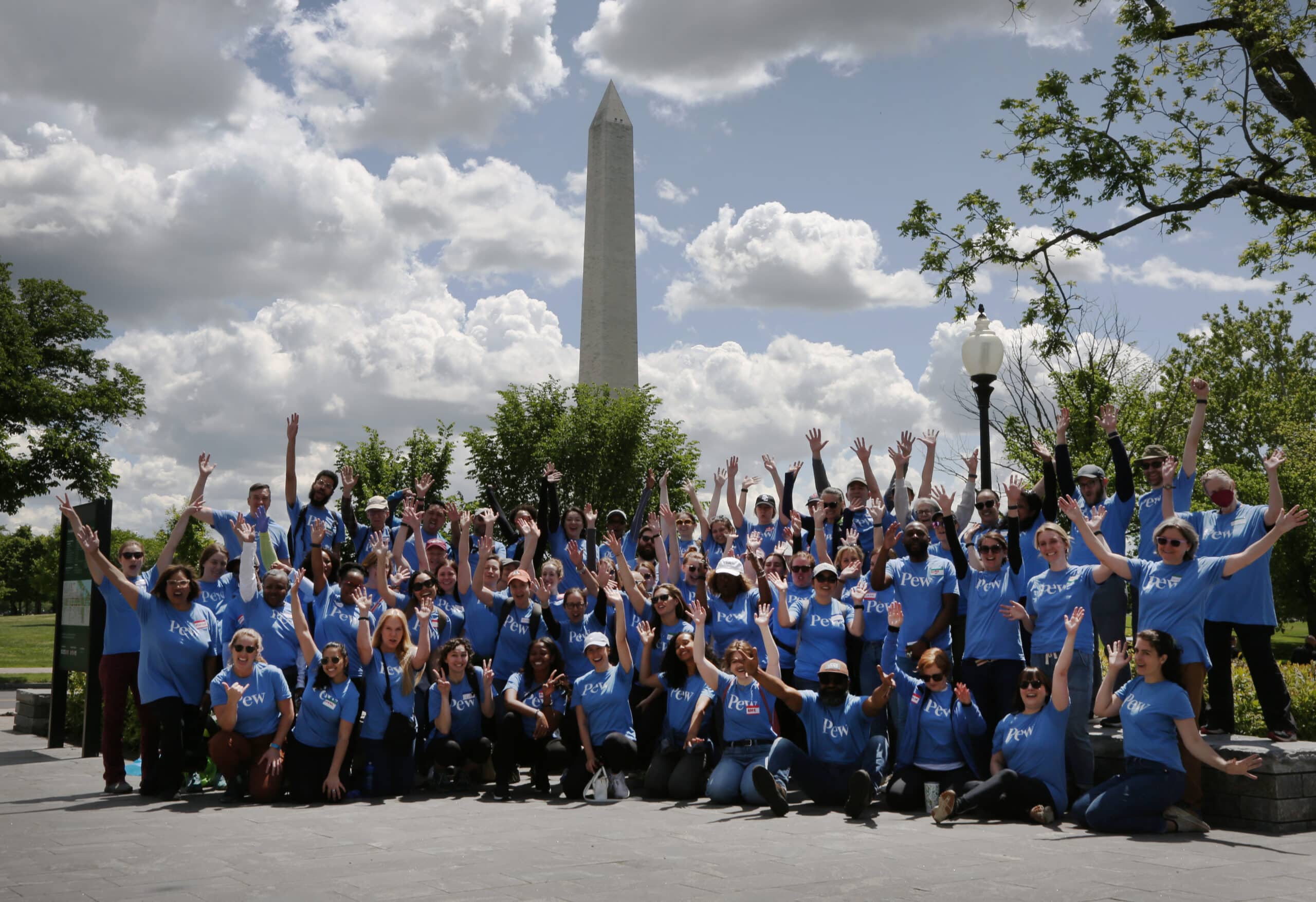 Nearly 100 Pew employees dressed in blue t-shirts raise their arms as they pose as a group in front of the Washington Monument.